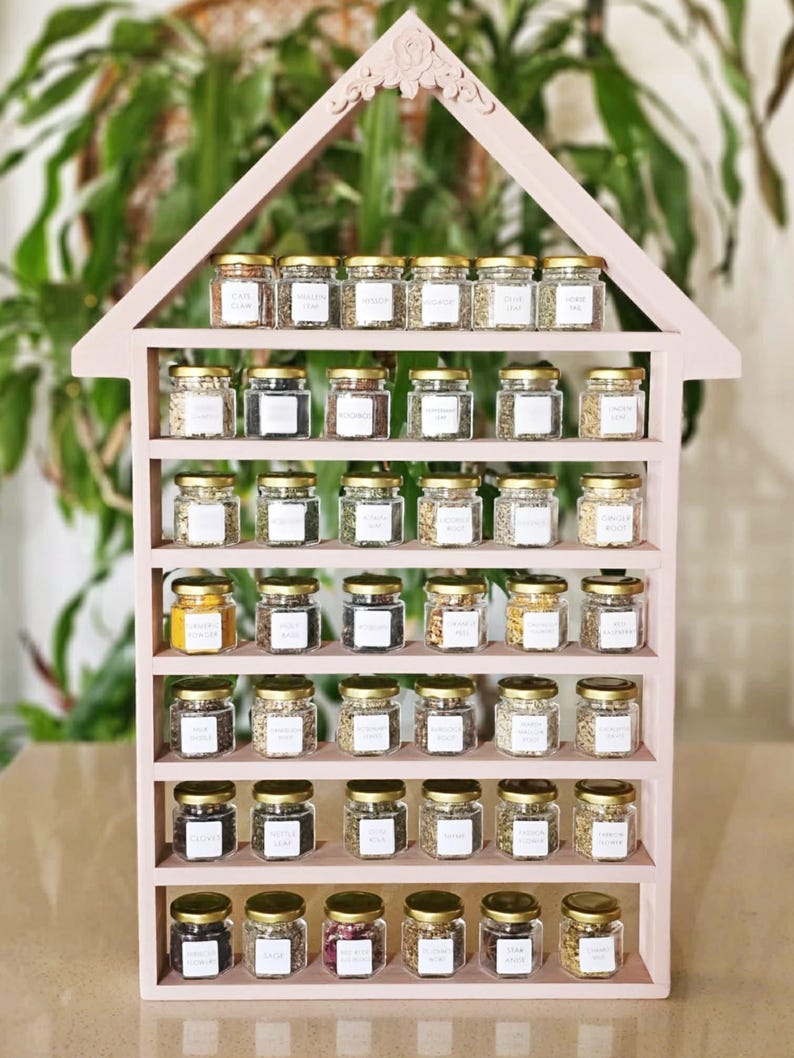 May include: A pink, house-shaped spice rack filled with small glass jars. Each jar has a gold lid and a white label. The rack is made of wood and has multiple shelves. The jars contain various spices, herbs, and seasonings.