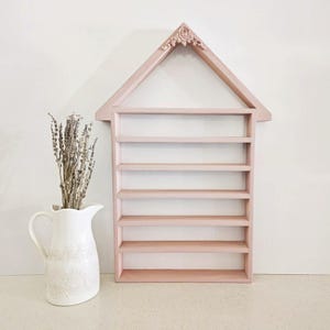 May include: A light pink wooden shelf in the shape of a house with six shelves. The top of the house has a decorative floral design. A white pitcher with dried lavender is to the left of the shelf.