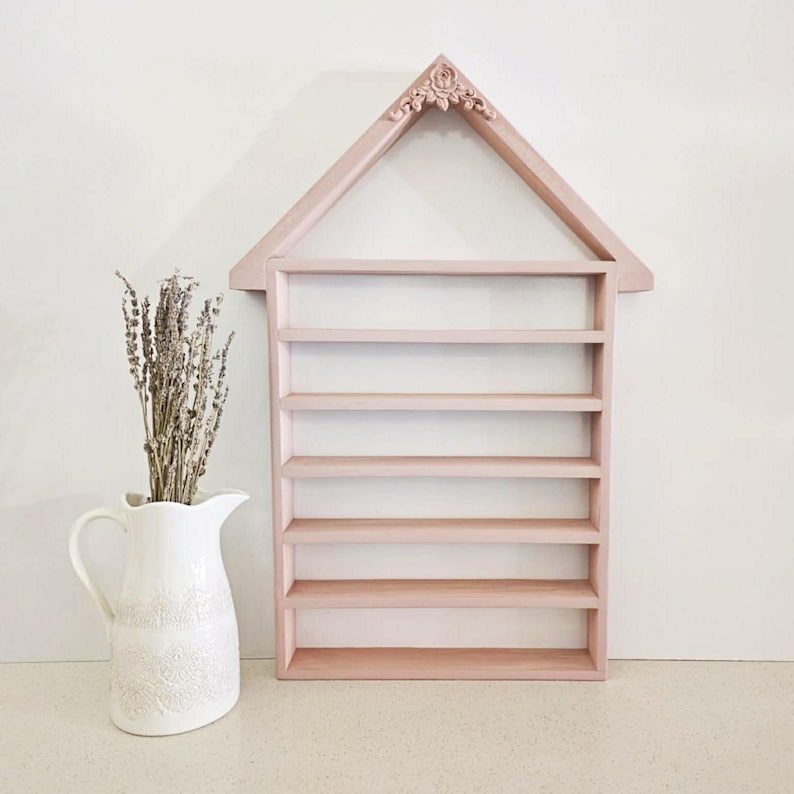 May include: A light pink wooden shelf in the shape of a house with six shelves. The top of the house has a decorative floral design. A white pitcher with dried lavender is to the left of the shelf.