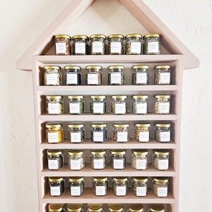May include: A pink, house-shaped spice rack filled with small glass jars. Each jar has a gold lid and a white label. The rack is mounted on a white wall. The jars contain various herbs and spices.