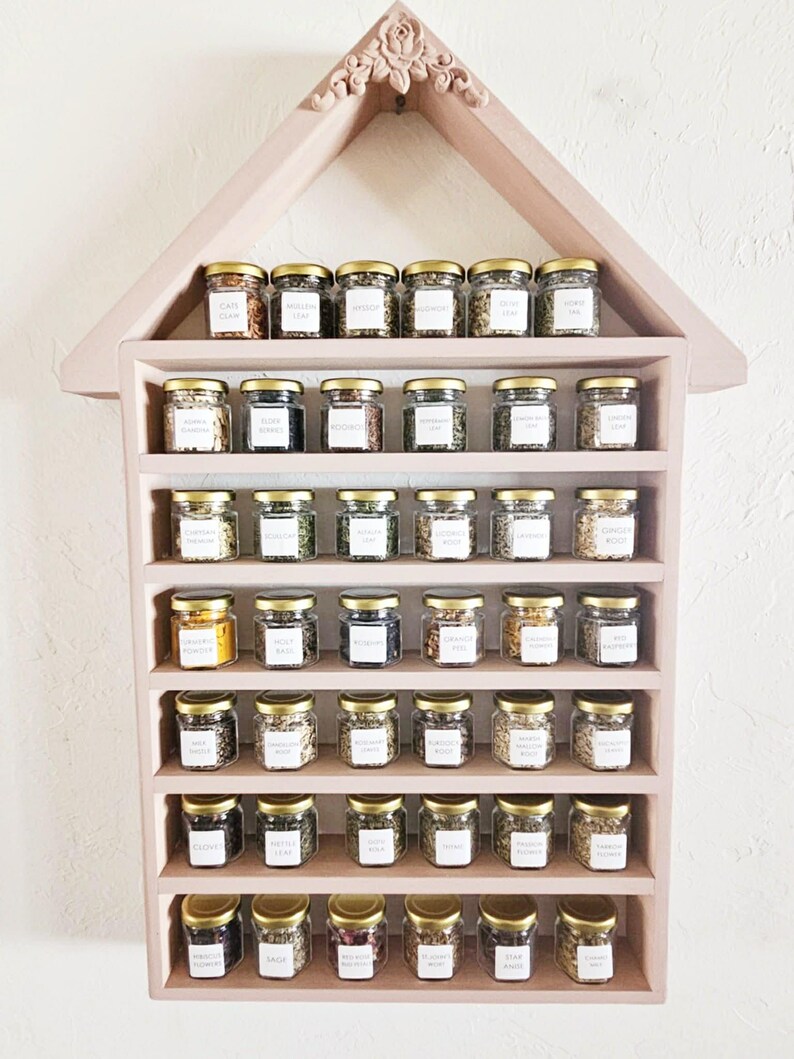 May include: A pink, house-shaped spice rack filled with small glass jars. Each jar has a gold lid and a white label. The rack is mounted on a white wall. The jars contain various herbs and spices.