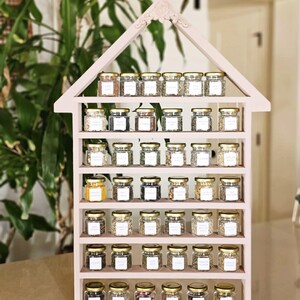 May include: A pink, house-shaped spice rack filled with small glass jars with gold lids. Each jar contains a different spice and has a white label. The rack has multiple shelves and is displayed against a backdrop of green plants.