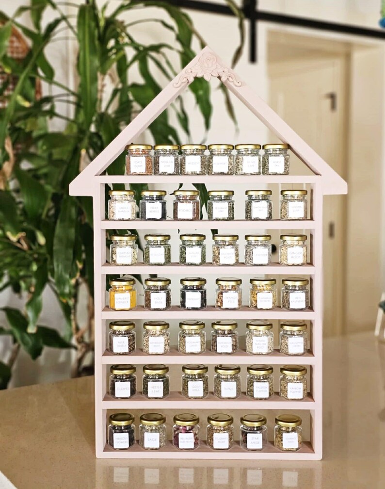 May include: A pink, house-shaped spice rack filled with small glass jars with gold lids. Each jar contains a different spice and has a white label. The rack has multiple shelves and is displayed against a backdrop of green plants.