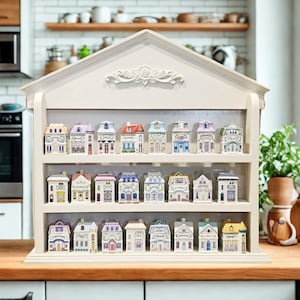 May include: A white wooden shelf unit shaped like a house, displaying miniature ceramic houses. The houses are painted in various colors with detailed architectural features. The shelf is on a wooden countertop.