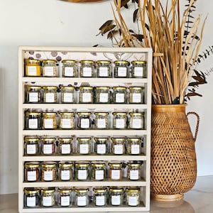 May include: A white wooden spice rack filled with small glass jars with gold lids. Each jar contains a different spice and has a white label. A woven vase with dried plants is to the right of the rack.