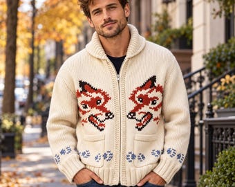 May include: A cream-colored, chunky knit zip-up cardigan featuring a shawl collar and two front pockets. The sweater displays a repeating red and black fox head pattern on the chest and blue paw print motifs along the ribbed cuffs and hem.
