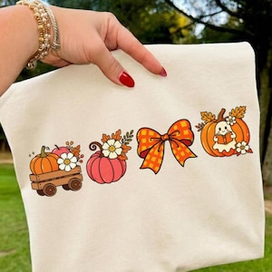 May include: A cream-colored sweatshirt featuring a graphic print of a wooden wagon carrying a pumpkin and flowers, a pink pumpkin with flowers, an orange checkered bow, and a pumpkin with a ghost reading a book and autumn leaves.