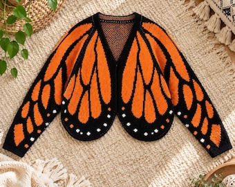 May include: A long-sleeved knit cardigan featuring a vibrant orange and black monarch butterfly wing pattern. The garment has a V-neckline, a black trim along the edges, and small white dot accents along the lower hem of the wing design.