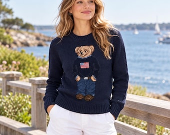 May include: A navy blue long-sleeve knit sweater featuring a central graphic of a teddy bear wearing a dark sweater with an American flag design, blue jeans, and brown shoes, paired with plain white shorts.