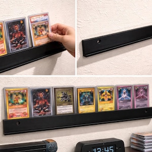 May include: Black metal display shelf holding several trading cards in protective cases. The cards feature various colorful illustrations and designs. The shelf is mounted on a white wall. A Pikachu figurine and a digital clock are visible.