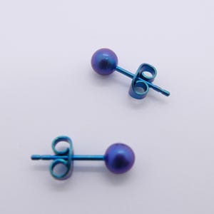 May include: Two blue stud earrings with a small, round, purple-blue bead at the end of each post. The earrings are set against a white background.