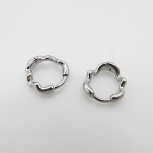 Op de afbeelding: Een paar kleine zilverkleurige oorringen met een onregelmatig, organisch en golvend randontwerp. De oorbellen zijn voorzien van een scharniersluiting en een gepolijste metalen afwerking, in een cirkelvorm met ongelijkmatige, getextureerde contouren.