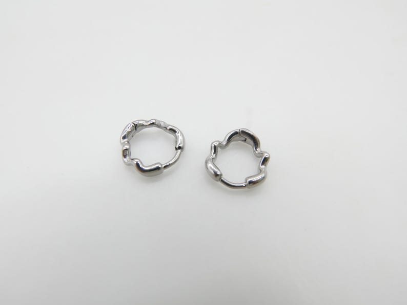 Op de afbeelding: Een paar kleine zilverkleurige oorringen met een onregelmatig, organisch en golvend randontwerp. De oorbellen zijn voorzien van een scharniersluiting en een gepolijste metalen afwerking, in een cirkelvorm met ongelijkmatige, getextureerde contouren.
