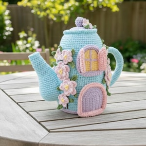 May include: A light blue crocheted teapot with a whimsical fairy house design. The teapot features a spout, handle, and lid, adorned with pink and yellow crocheted flowers and a door and window. The teapot is sitting on a wooden table.