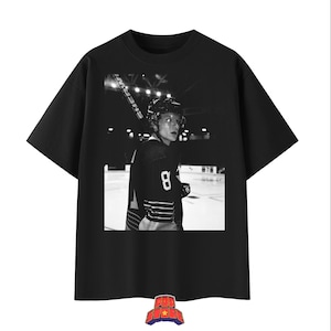 May include: Black t-shirt featuring a black and white image of a hockey player in uniform, holding a hockey stick. The player is wearing a helmet and the number 8 is visible on the jersey. The shirt has a crew neck.