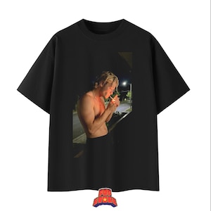 Puede incluir: Una camiseta negra de manga corta con un estampado gráfico rectangular en el pecho. El estampado muestra a una persona sin camisa y con cabello rubio encendiendo un cigarrillo por la noche, iluminada por un brillo cálido. Cuello redondo y corte recto.