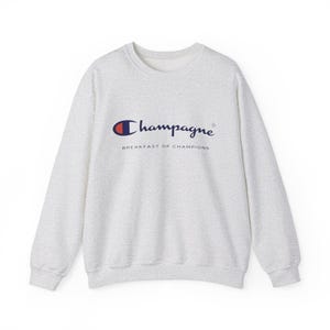 May include: A light gray sweatshirt with the word "Champagne" in a navy blue script, above "BREAKFAST OF CHAMPIONS" in a smaller font. The "Champagne" text is above a Champion logo in red, white, and blue.
