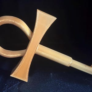 May include: A gold-colored object shaped like an ankh symbol combined with a key. The item features a rounded loop at the top, a vertical crossbar, and a straight shaft extending to a small, rectangular bit at the end, showing a textured surface.