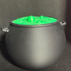 May include: A matte black cauldron with two small side handles, featuring a bright green, textured liquid surface that appears to be bubbling with small, raised circular protrusions across the top of the vessel.
