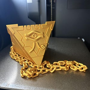 Peut inclure: Une boîte triangulaire dorée avec un symbole d'œil et une chaîne. La boîte a une forme de pyramide avec un motif détaillé sur le devant. Une chaîne dorée est drapée autour de la base de la boîte. Il s'agit probablement d'un objet de collection ou décoratif.