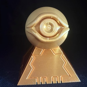 Peut inclure: Un objet imprimé en 3D de couleur dorée présentant une partie supérieure sphérique avec un motif d'œil sculpté au centre. La sphère repose sur une base pyramidale qui affiche des motifs de lignes géométriques en creux sur sa face avant.