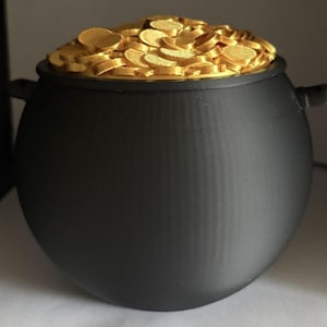 Puede incluir: Un caldero negro mate con dos pequeñas asas laterales, lleno hasta el borde con un montón de monedas circulares brillantes de color dorado. El recipiente tiene un cuerpo redondeado y una textura suave y uniforme que contrasta con las monedas.