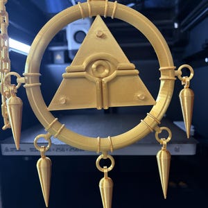 Könnte beinhalten: Goldfarbener Anhänger mit kreisförmigem Rahmen, zentralem Pyramiden-Design und Augensymbol. Der Anhänger hat konisch geformte Anhänger am Rahmen und wird von einer Kette gehalten.