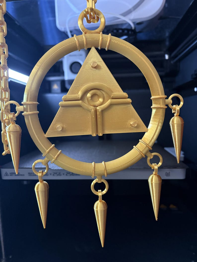 May include: A gold-colored pendant featuring a circular frame with a central pyramid design and an eye symbol. The pendant has several cone-shaped dangles attached to the frame, and is suspended by a chain.