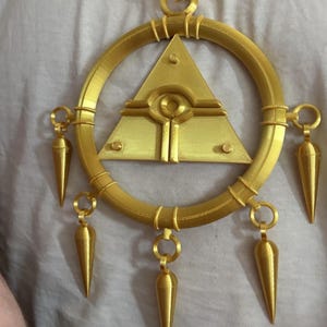 May include: Gold-colored pendant necklace featuring a central triangle with an eye design, surrounded by a circular frame. The pendant has several cone-shaped dangles. The necklace is made of a gold-colored material.