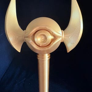 Könnte beinhalten: Ein goldfarbener Stab mit einem zentralen kugelförmigen Kopf und einem skulptierten Augendesign. Zwei geschwungene, flügelartige Vorsprünge ragen seitlich aus der Kugel heraus. Das gesamte Objekt weist eine 3D-gedruckte Textur mit feinen Linien auf.
