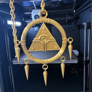 May include: A gold-colored, 3D-printed pendant with a circular frame, a central triangle with an eye design, and dangling cone-shaped ornaments. The pendant is suspended by a chain.