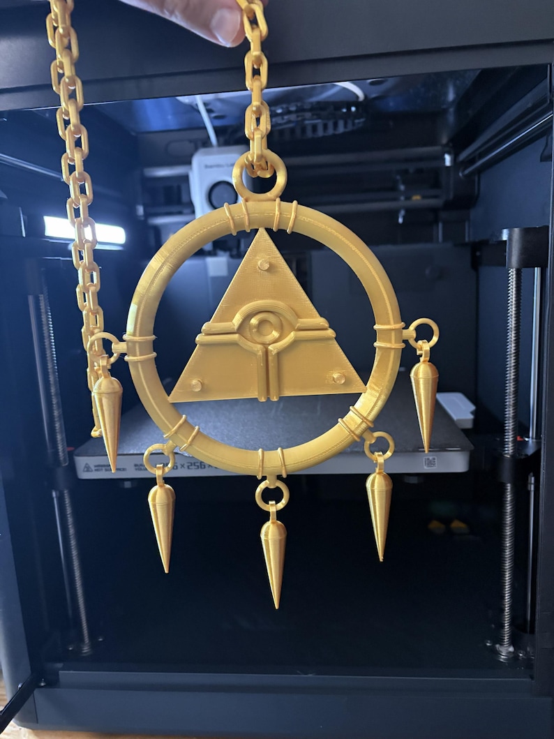 May include: A gold-colored, 3D-printed pendant with a circular frame, a central triangle with an eye design, and dangling cone-shaped ornaments. The pendant is suspended by a chain.