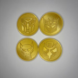 May include: Four gold-coloured coins with embossed designs. Two coins feature the Autobot symbol, while the other two display different character emblems. The coins are circular and have a metallic appearance.