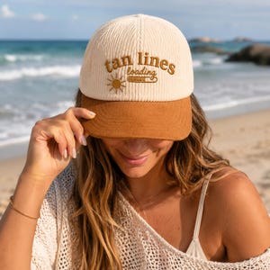 May include: A cream-colored corduroy baseball cap with a brown brim. The cap features the embroidered text "tan lines loading" with a sun graphic. The hat is worn on a beach.