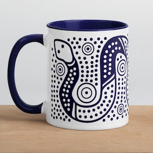 Peut inclure: Un mug en céramique blanche avec un intérieur et une anse bleu foncé. L'extérieur présente un motif graphique bleu foncé représentant un serpent composé de points et de cercles concentriques, couvrant le côté du récipient cylindrique.