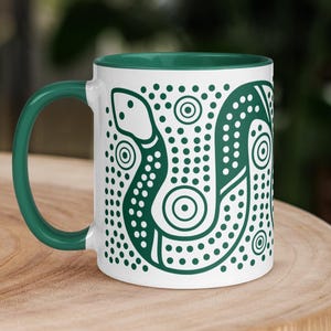 May include: A white ceramic mug with a dark green interior and handle. The exterior features a dark green graphic design of a snake composed of dots and concentric circles, set against a background of scattered dark green dots.