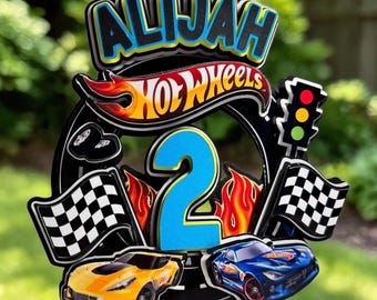 May include: A birthday cake topper featuring the name "ALIJAH" in blue and yellow, the Hot Wheels logo, a blue number 2, race flags, a traffic light, and two toy cars. The topper is black with colorful accents.
