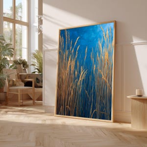 Puede incluir: Una impresión artística enmarcada que presenta altas hierbas de color marrón dorado contra un fondo azul profundo. La obra de arte se muestra en un marco de madera clara, apoyada contra una pared blanca. La pieza evoca una sensación de naturaleza y tranquilidad.