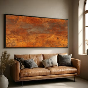 May include: A large framed abstract art piece with a rusty orange and brown color palette. The artwork hangs above a brown leather sofa with decorative pillows. The room has a neutral color scheme.
