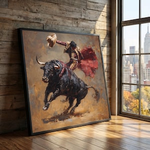May include: A framed painting depicts a bullfight scene. A matador in a gold and red suit is on a black bull. The bull is running, and the matador is holding his hat. The background is a warm, neutral color.