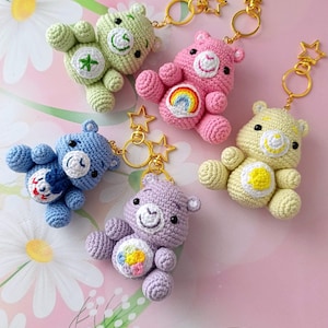 May include: Five colorful crochet bear keychains. Each bear is a different color: green, pink, blue, purple, and yellow. Each bear has a unique embroidered design on its belly and a gold star keychain attachment.