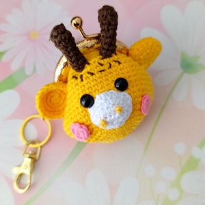 May include: A handmade yellow crochet coin purse shaped like a giraffe's head. It features black eyes, a white muzzle, pink cheeks, and brown antlers. The purse has a gold-colored clasp and a matching keychain.