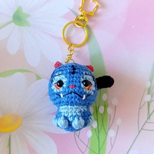 May include: A handmade blue crochet keychain featuring a cartoon-like creature with red accents and black details. The keychain has a gold-colored ring and a cat-shaped clasp.
