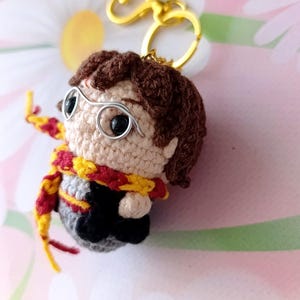 May include: A handmade crochet keychain featuring a character with brown hair, glasses, and a red and yellow scarf. The keychain has a gold-colored clasp. The character is wearing a gray and black outfit. The background is a soft pink and white floral pattern.