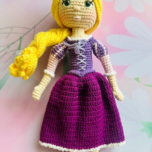 May include: A handmade crochet doll with yellow hair and a purple dress. The doll has a yellow crocheted head and long braid, green eyes, and a purple bodice with white lacing. The skirt is a deep purple with a white trim, and the doll wears purple shoes.