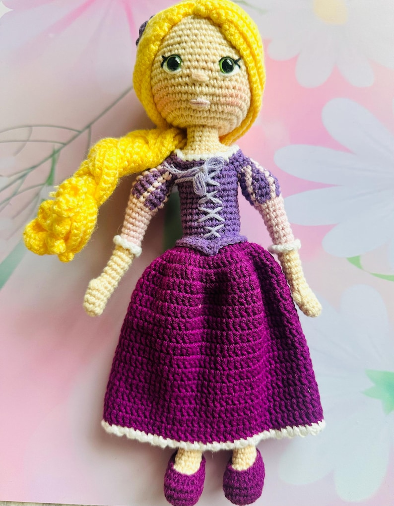 May include: A handmade crochet doll with yellow hair and a purple dress. The doll has a yellow crocheted head and long braid, green eyes, and a purple bodice with white lacing. The skirt is a deep purple with a white trim, and the doll wears purple shoes.