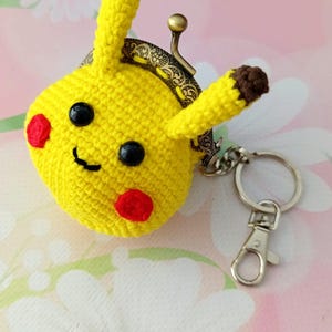 May include: A yellow crocheted coin purse shaped like a cartoon character with black eyes, red cheeks, and brown-tipped ears. The purse has a metal clasp and a keychain attachment.