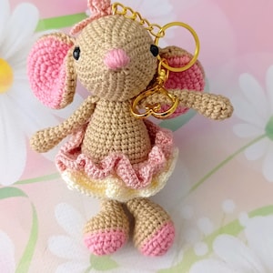 May include: A crocheted mouse keychain in tan, pink, and yellow. The mouse has pink ears, a pink nose, and a pink bow. It wears a pink and yellow tutu. The keychain has a gold-colored chain and clasp.
