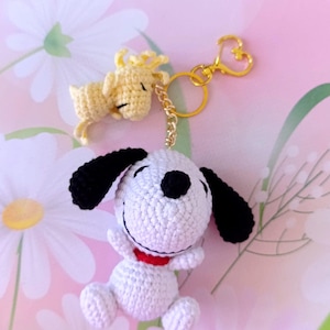 May include: Crocheted Snoopy and Woodstock keychain. The Snoopy figure is white with black ears and a red collar. Woodstock is yellow. Both are attached to a gold-colored heart-shaped clasp.