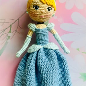 May include: A handmade crochet doll depicting a princess in a blue dress. The doll has yellow hair styled in a bun, blue eyes, and a light skin tone. The dress features a detailed crochet pattern and light blue accents.